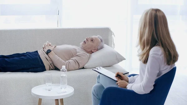 Blonde psychotherapist holding pen near clipboard and listening middle aged man lying on couch in consulting room — Fotografia de Stock