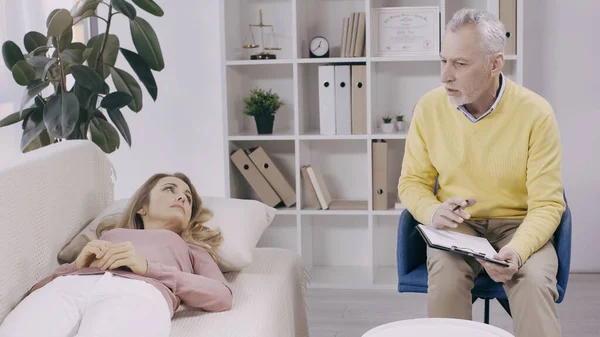 Blonde woman lying on couch and looking at middle aged psychotherapist with clipboard — Stock Photo