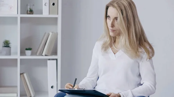 Concentrated psychologists writing on clipboard and looking away in consulting room — Stock Photo