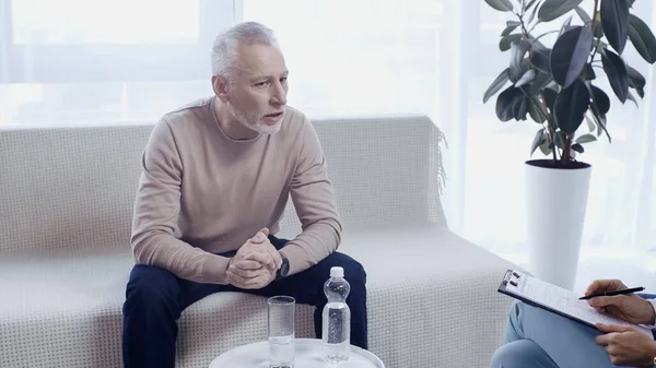 Mann mittleren Alters sitzt mit geballten Händen auf Sofa neben Psychotherapeut — Stockfoto