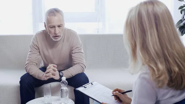 Middle aged man with clenched hands sitting on sofa near blonde psychotherapist — Foto stock