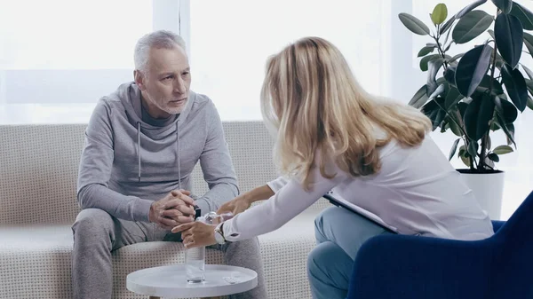 Middle aged man in sportswear with clenched hands sitting on sofa near psychotherapist pouring water in glass — Stockfoto