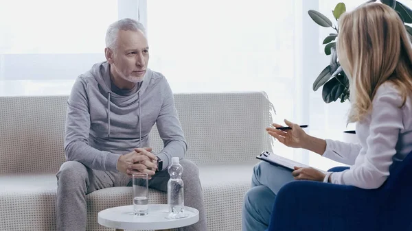 Hombre de mediana edad en ropa deportiva con las manos apretadas sentado en el sofá cerca de psicoterapeuta en la sala de consulta - foto de stock