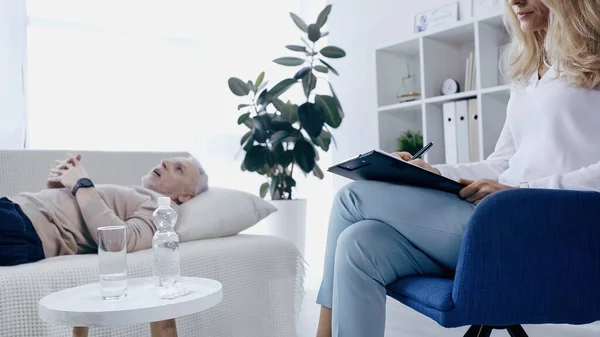 Psicoterapeuta escribir en portapapeles cerca borrosa hombre de mediana edad con las manos apretadas acostado en el sofá - foto de stock