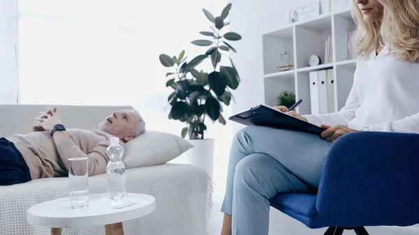 Psychotherapist writing on clipboard near blurred middle aged man with clenched hands lying on couch — стоковое фото