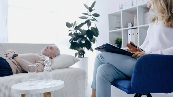 Psicoterapeuta sosteniendo la pluma cerca del portapapeles y hablando con un hombre de mediana edad borroso con las manos apretadas acostado en el sofá - foto de stock