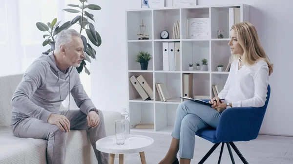 Middle aged man in sportswear sitting on sofa near blonde psychologist in consulting room — Stockfoto