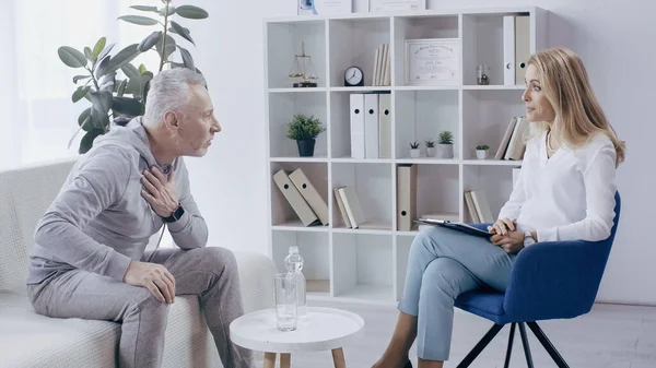 Hombre de mediana edad en ropa deportiva sentado en el sofá y hablando con la psicóloga rubia en la sala de consulta - foto de stock