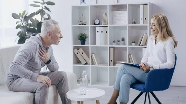 Middle aged man in sportswear sitting on sofa and talking to cheerful psychologist in consulting room — Stock Photo