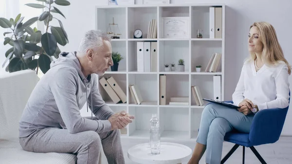 Homem de meia idade em sportswear sentado no sofá e falando com psicólogo sorridente em consultório — Fotografia de Stock
