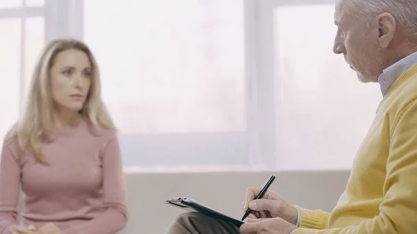 Psicoterapeuta de mediana edad sosteniendo la pluma cerca del portapapeles y hablando con el paciente borroso en la sala de consulta - foto de stock