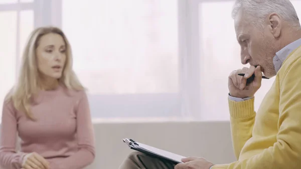 Thoughtful middle aged psychotherapist holding pen near clipboard and listening blurred patient in consulting room — Stock Photo