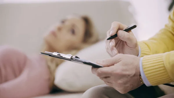 Закрыть обзор зрелого психотерапевта, держащего планшет и ручку рядом с размытым пациентом — стоковое фото