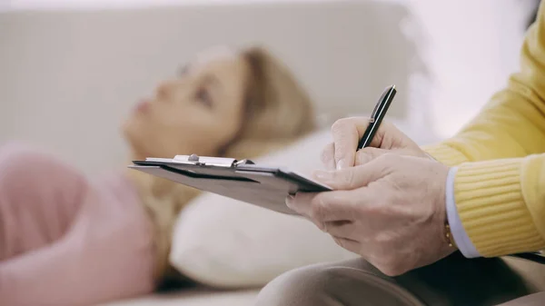 Close up of mature psychotherapist writing on clipboard near blurred patient — Stock Photo