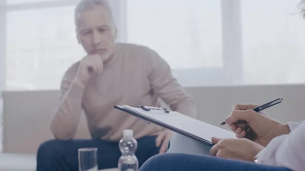 Psychotherapist writing on clipboard near pensive blurred man in consulting room — стоковое фото