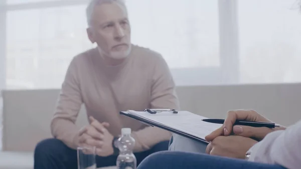 Psychologue tenant stylo et presse-papiers près de l'homme flou dans la salle de consultation — Photo de stock