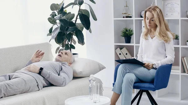 Psicoterapeuta escribir en portapapeles cerca de hombre de mediana edad acostado en el sofá durante la consulta - foto de stock