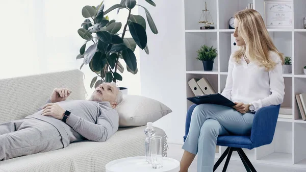 Psychotherapist holding clipboard near middle aged man lying on couch and talking during consultation — Foto stock