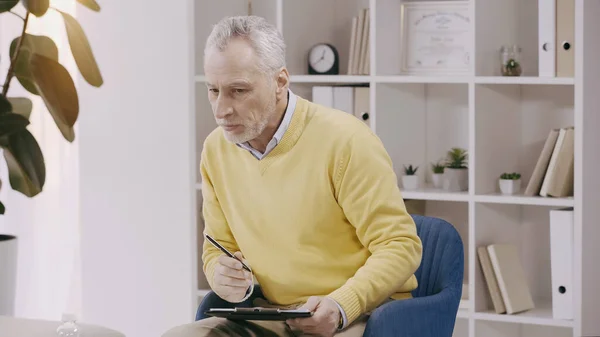 Attentive mature psychologist listening and holding clipboard during consultation — Stockfoto