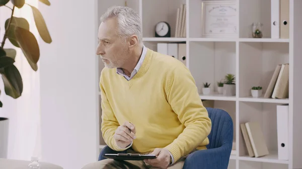 Mature psychologist listening and holding clipboard during consultation — стоковое фото