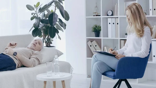 Rubia psicóloga escribiendo en portapapeles cerca de hombre de mediana edad acostado en el sofá y hablando durante la consulta - foto de stock