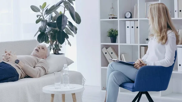 Rubia psicólogo sosteniendo portapapeles cerca de mediana edad hombre acostado en el sofá y hablando durante la consulta - foto de stock