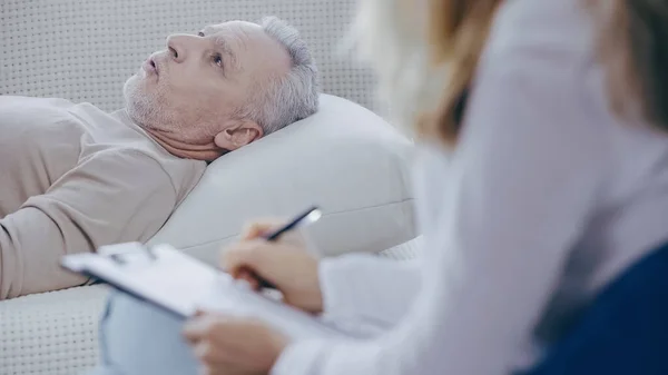 Middle aged man lying on couch and talking during consultation with blurred psychologist holding clipboard — стоковое фото
