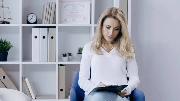 Rubia psicólogos escribiendo en portapapeles cerca de rack en la sala de consulta - foto de stock