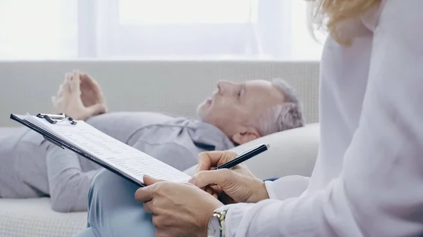Psicologo tenendo appunti vicino sfocato uomo di mezza età sdraiato sul divano durante la consultazione — Foto stock