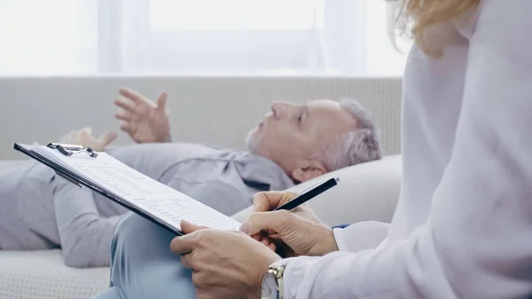 Psychologe hält Klemmbrett und Stift in der Nähe eines verschwommenen Mannes mittleren Alters, der während der Beratung auf dem Sofa liegt — Stockfoto
