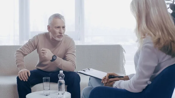 Blonde psychologist holding pen and clipboard near man talking in consulting room — Foto stock