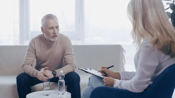 Чоловік середнього віку розмовляє з блондинкою-психологом, тримаючи ручку і буфер обміну в консалтинговій кімнаті — стокове фото