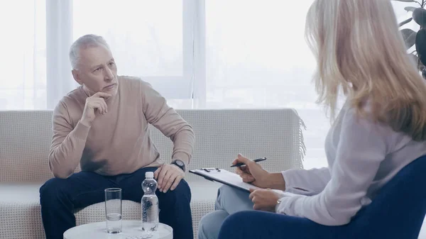 Blonde psychologist holding clipboard near pensive middle aged man in consulting room — Fotografia de Stock