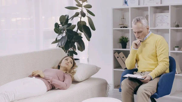 Blonde woman lying on couch near thoughtful middle aged psychotherapist with clipboard — Fotografia de Stock