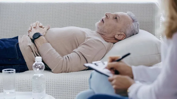 Blurred psychologist writing on clipboard near pensive middle aged man lying on sofa during therapy — Fotografia de Stock