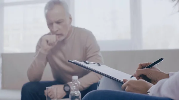 Psychologist holding clipboard near worried middle aged man sitting on sofa during therapy — Foto stock