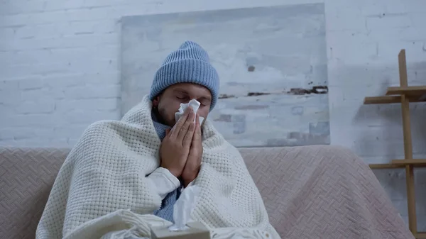 Hombre enfermo en gorro caliente y manta estornudando en servilleta de papel mientras está sentado en el sofá - foto de stock