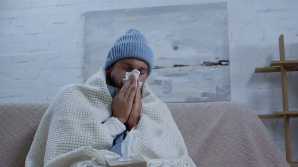Sick man in warm beanie and blanket sneezing in paper napkin while sitting on sofa — Stock Photo