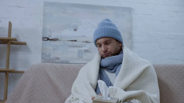 Sick man with pack of paper napkins sitting on sofa in warm beanie, scarf and blanket — Foto stock