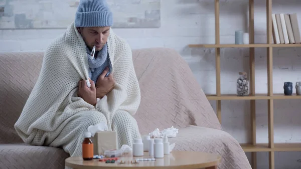 Хворий чоловік вимірює температуру сидячи на дивані в теплій ковдрі і бані біля ліків — стокове фото