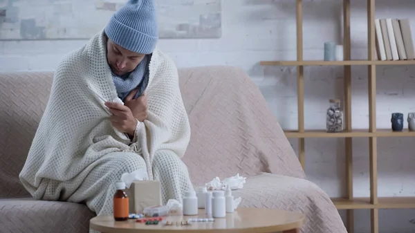 Хворий чоловік сидить на дивані в теплій ковдрі і капелюсі біля столу з паперовими серветками і ліками — стокове фото