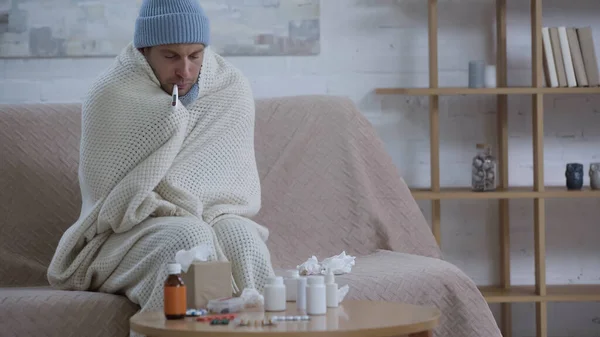 Hombre enfermo en sombrero caliente y gorro de medir la temperatura cerca de la mesa con medicamentos - foto de stock