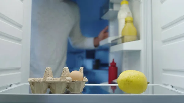 Cropped view of blurred man near eggs, lemon and sauces in fridge — стоковое фото