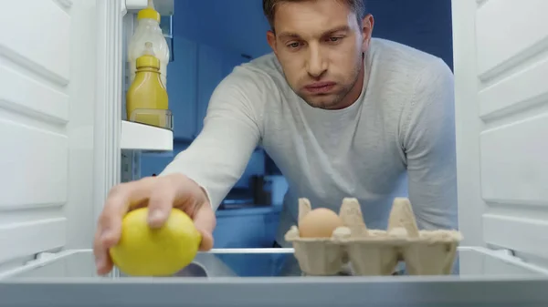 Незадоволений чоловік надуває щоки, приймаючи лимон з холодильника — стокове фото