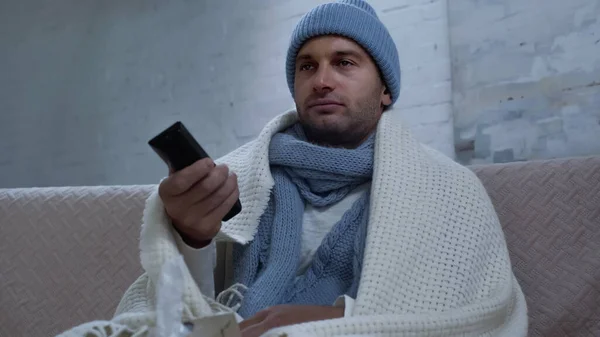 Sick man in warm beanie, scarf and blanket clicking channels while watching tv — Foto stock