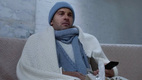 Diseased man in warm hat, scarf and blanket watching tv on couch at home — Photo de stock