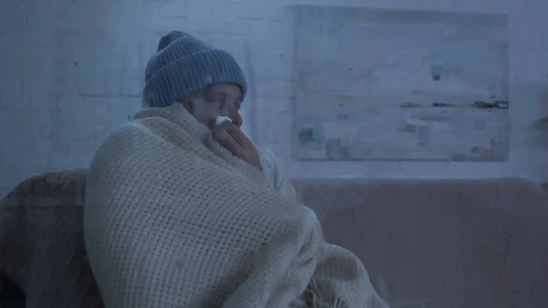 Diseased man in warm hat and blanket sneezing in paper napkin on couch at home — Fotografia de Stock