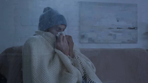 Sick man sitting on couch in warm beanie and blanket and sneezing in paper napkin — Fotografia de Stock