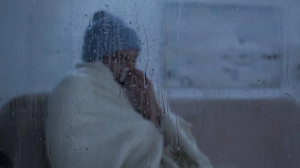 Hombre enfermo en sombrero caliente y manta sentado en casa cerca de la ventana con gotas de lluvia - foto de stock