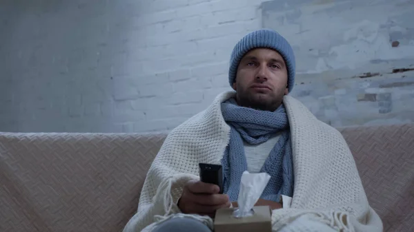 Homme malade avec télécommande tv et serviettes en papier assis sur le canapé en bonnet chaud et couverture — Photo de stock